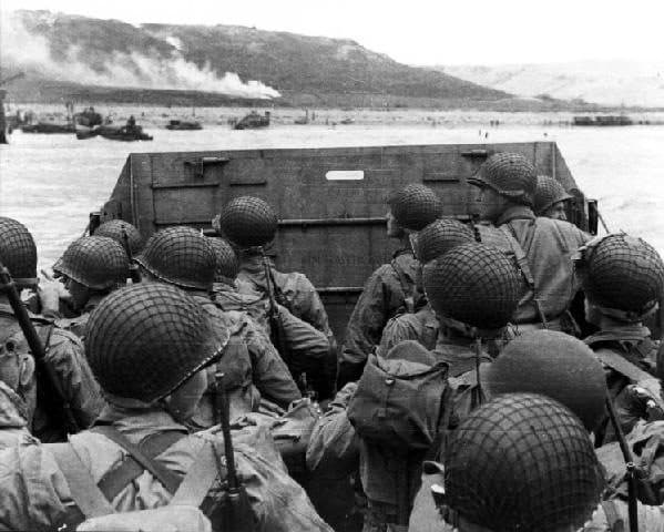 Helmeted troops packed into a landing craft heading toward a smoking shoreline